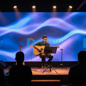Một nghệ sĩ guitar người Việt biểu diễn trên một sân khấu nhỏ trong một quán cafe acoustic ấm cúng, low-lit. Màn hình LED Indoor P2.5 đóng vai trò làm backdrop động, hiển thị họa tiết ánh sáng xanh tím.