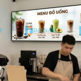 Một menu điện tử kỹ thuật số sắc nét, hiển thị hình ảnh hấp dẫn về các loại đồ uống Việt Nam (cà phê sữa đá, sinh tố bơ) và giá cả, được lắp đặt phía trên quầy pha chế tại một quán cafe hiện đại. Một barista đang làm việc phía dưới.