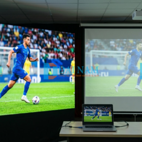 Bức ảnh so sánh thực tế giữa màn hình LED (bên trái) hiển thị hình ảnh bóng đá rực rỡ, độ tương phản cao và máy chiếu (bên phải) bị mờ, nhạt màu dưới ánh sáng đèn hội trường.